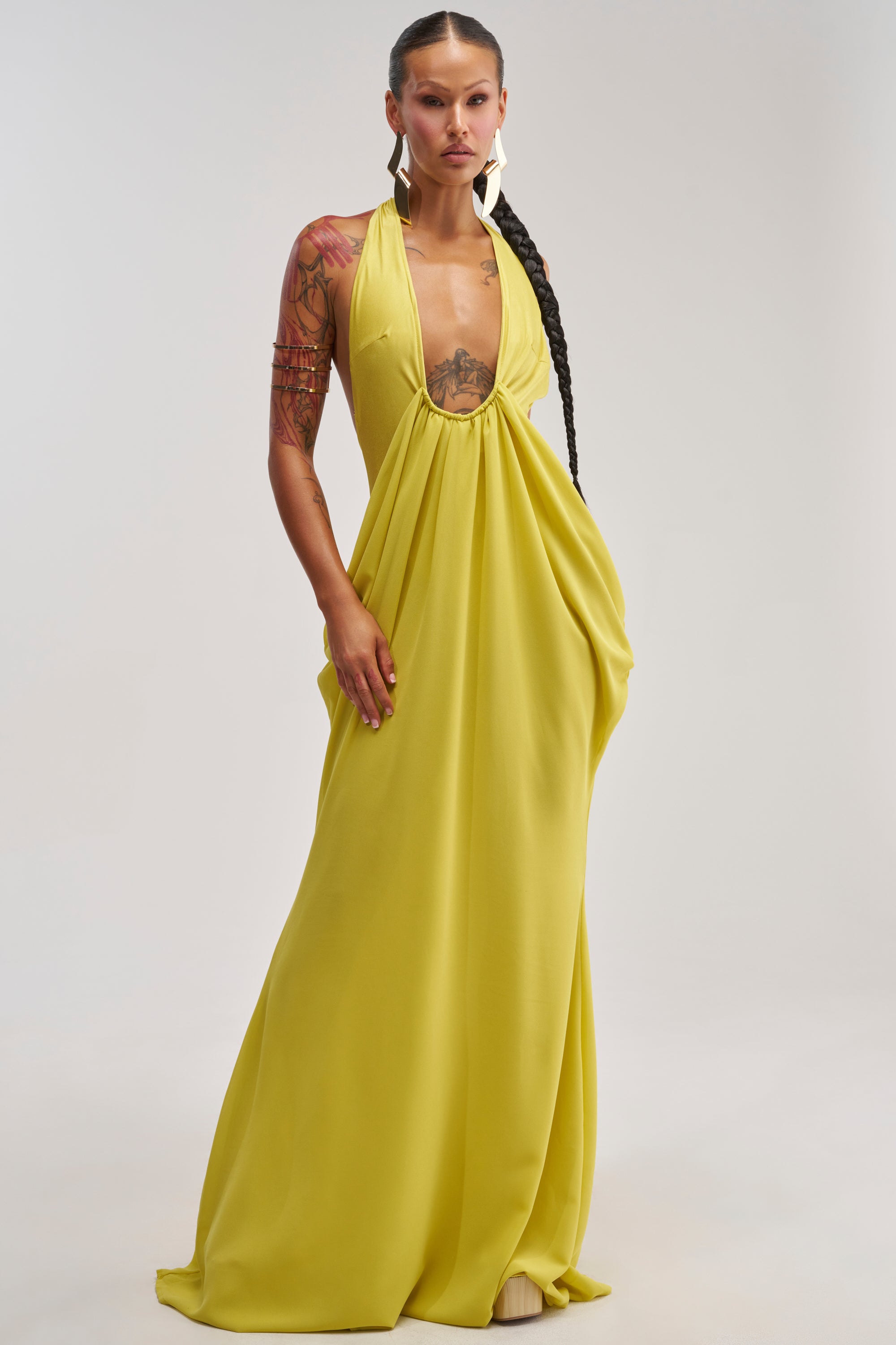 A person with long braided hair and tattoos wears the BORN TO SHINE SWOOP MAXI DRESS—a flowing, bright chartreuse maxi dress with a deep neckline, perfect for resort occasions—while standing against a plain light background.