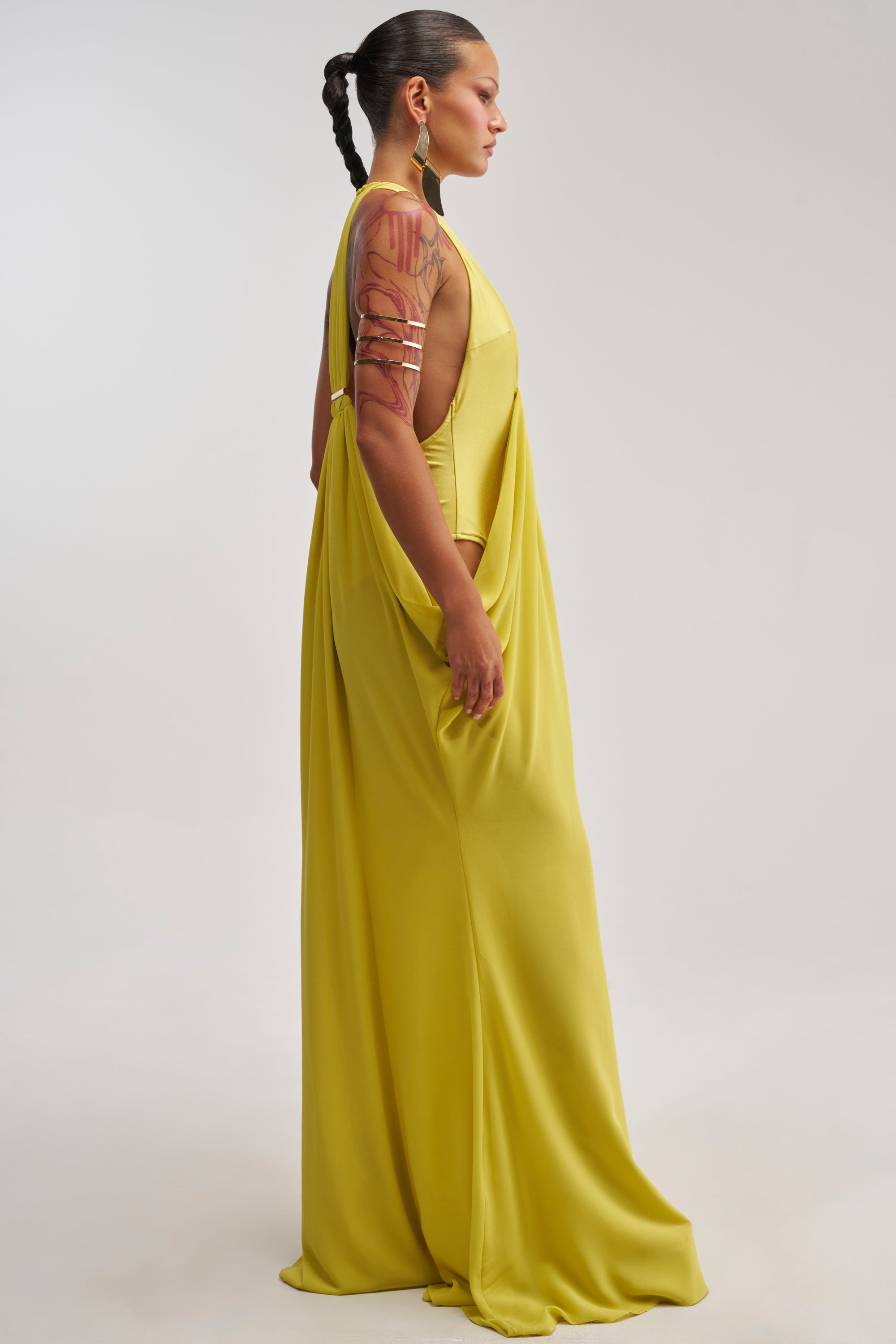 A woman with braided hair and an arm tattoo models the BORN TO SHINE SWOOP MAXI DRESS, a sleeveless, draped evening dress in a striking yellow hue. She stands in profile against a plain light background, embodying effortless elegance perfect for a resort occasion.