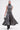 A woman in the DONT GIVE UP MOCK NECK KNIT MAXI DRESS, sleeveless and body-contouring with a flared hem, walks forward in black boots holding a small silver handbag against a plain white background.