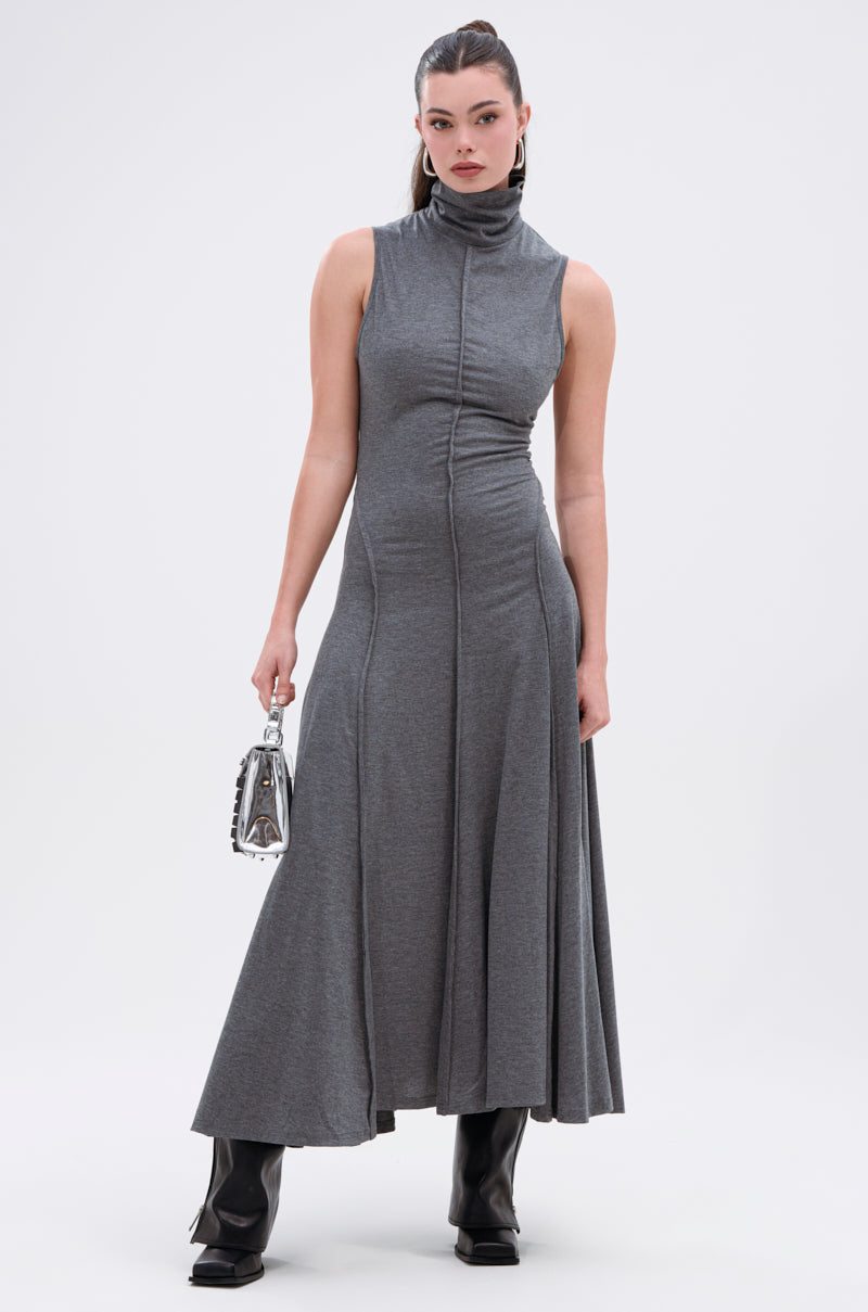 A woman models the DONT GIVE UP MOCK NECK KNIT MAXI DRESS—featuring a mock neck, flared hem, and body-contouring seams—with black boots and a metallic handbag, posed against a plain white background.