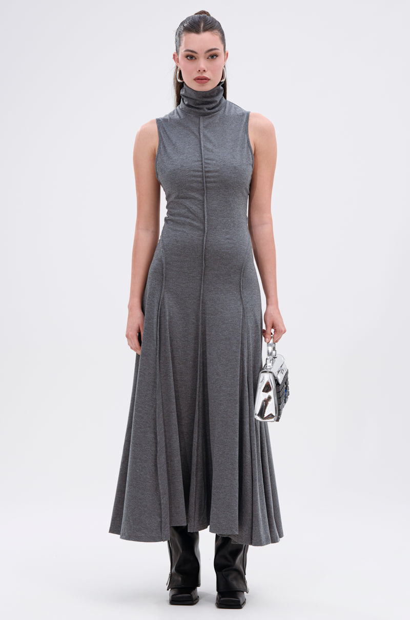 A woman faces forward in the DONT GIVE UP MOCK NECK KNIT MAXI DRESS with body-contouring seams and a flared hem. She holds a small silver handbag, wears black boots, and her hair is tied back in a sleek ponytail.