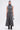 A woman faces forward in the DONT GIVE UP MOCK NECK KNIT MAXI DRESS with body-contouring seams and a flared hem. She holds a small silver handbag, wears black boots, and her hair is tied back in a sleek ponytail.