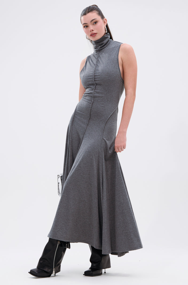 A woman wears the DONT GIVE UP MOCK NECK KNIT MAXI DRESS in grey, featuring body-contouring seams and a flared hem, paired with black heeled boots. She stands against a white background, holding a small silver handbag.