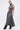 A woman wears the DONT GIVE UP MOCK NECK KNIT MAXI DRESS in grey, featuring body-contouring seams and a flared hem, paired with black heeled boots. She stands against a white background, holding a small silver handbag.