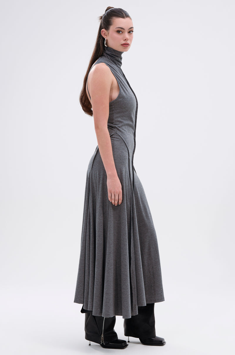 A woman in profile wears the DONT GIVE UP MOCK NECK KNIT MAXI DRESS with body-contouring seams, a flared hem, and black boots. Her long dark hair is half-up in a ponytail, set against a plain light background.