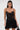 A woman with long dark hair and large hoop earrings wears the DORELLA LACE MINI DRESS, a sheer black floral lace piece with thin straps and a delicate sweetheart neckline, standing against a plain light background.