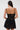 A woman with long dark hair and large hoop earrings is seen from the back wearing the DORELLA LACE MINI DRESS—black, skater silhouette, thin straps—against a plain light background.