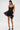A woman wearing the DORELLA LACE MINI DRESS with a sweetheart neckline, black heels, and a small black handbag stands against a plain white background.