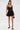 A woman stands against a white background wearing the DORELLA LACE MINI DRESS, black ankle-strap heels, large hoop earrings, and holding a small black handbag.