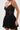 A woman wears the DORELLA LACE MINI DRESS, a sheer black lace mini dress with thin straps and a sweetheart neckline, standing against a plain light background and holding a small black clutch in her right hand.
