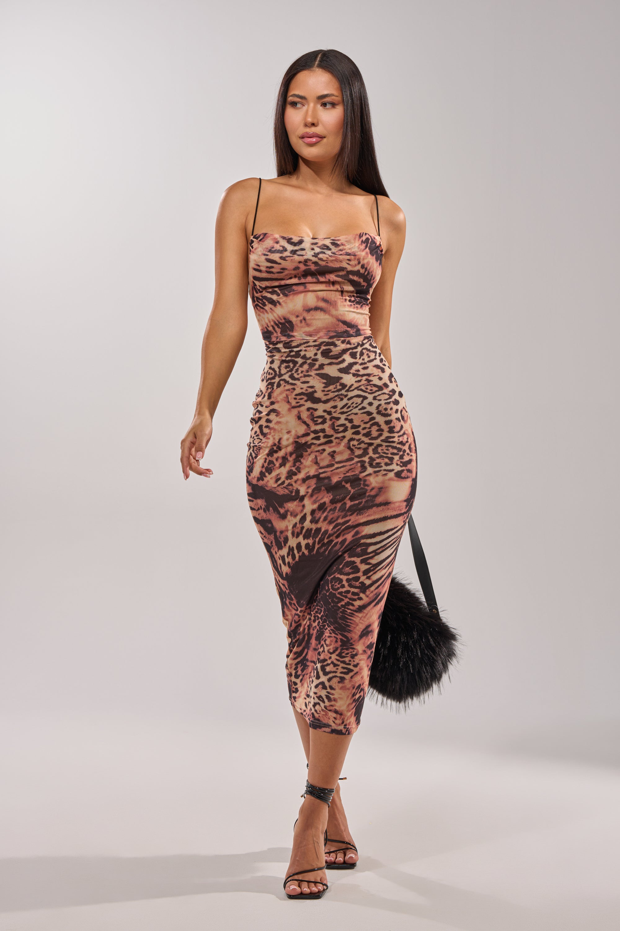 A woman wearing the CLAWS OUT LEOPARD MIDI SLIP DRESS with thin straps, black high-heeled sandals, and holding a black faux fur handbag stands against a plain light background. 0