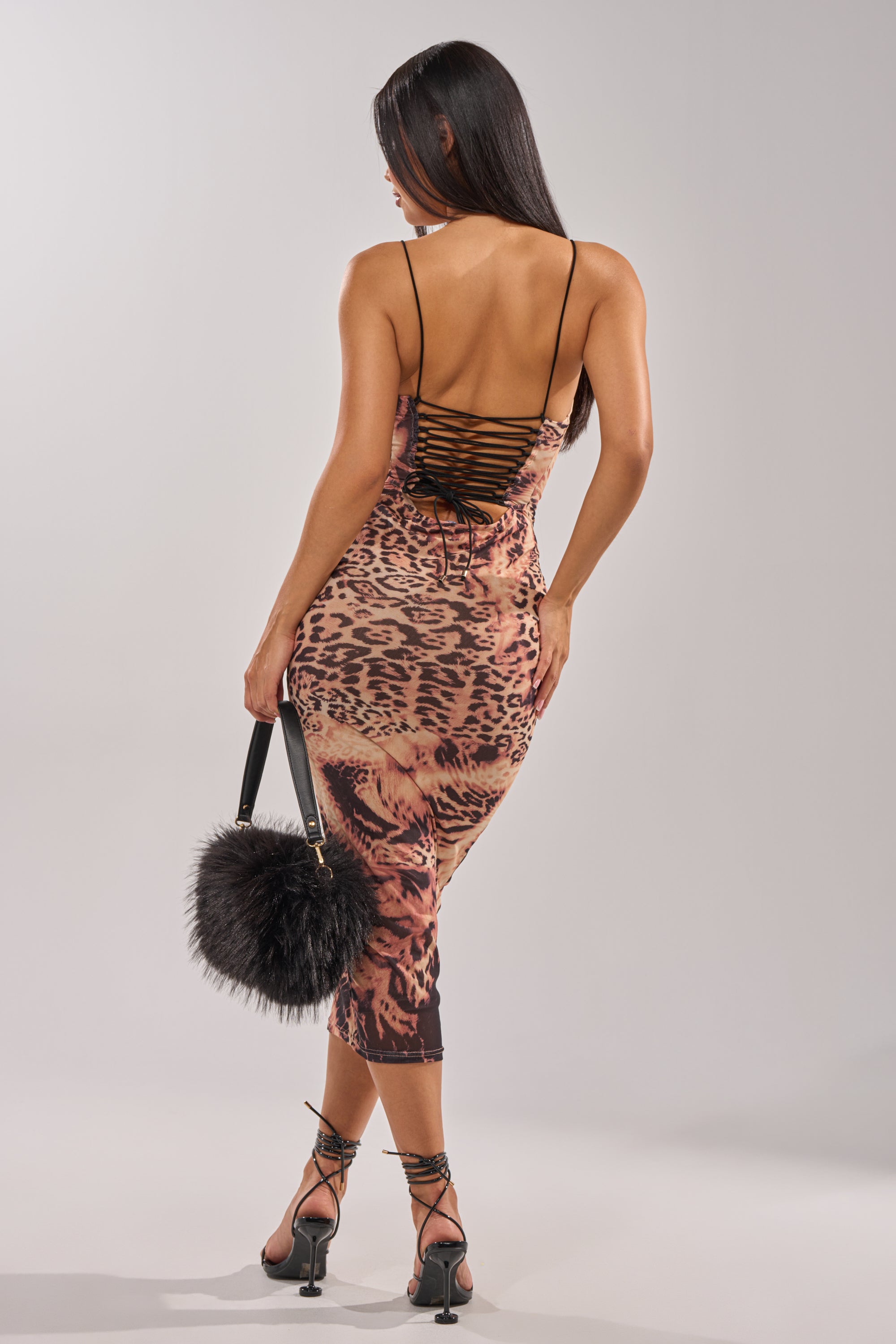 A woman faces away in the CLAWS OUT LEOPARD MIDI SLIP DRESS, a form-fitting slip with thin straps and lace-up detail. She accessorizes with a black fluffy handbag and strappy high heels. 3