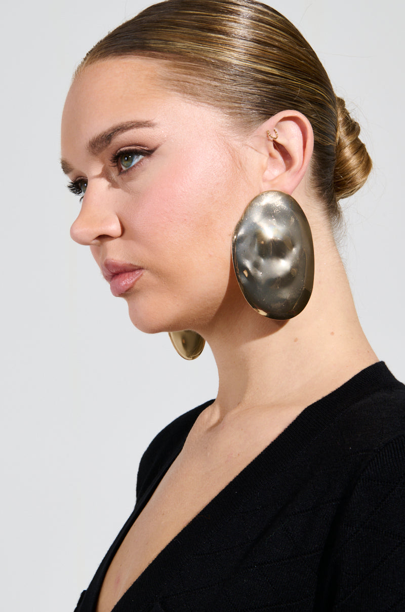 A person with slicked-back hair wears DIDI STATEMENT EARRINGS, oversized metallic discs with hammered detailing and a small stud. They're dressed in black and gazing sideways against a plain background.