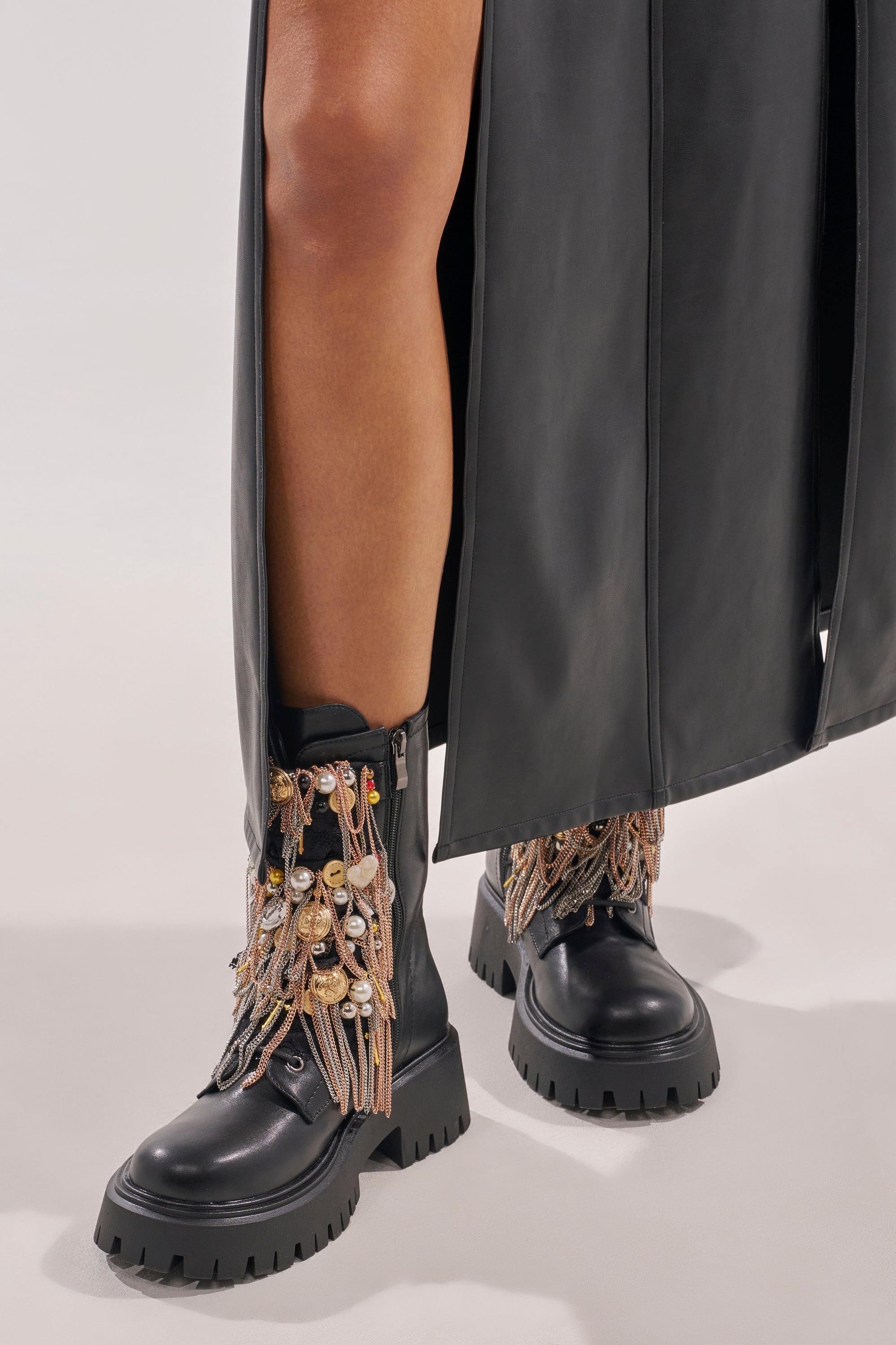 A person wears a long black leather skirt with AZALEA WANG MEGUMI BLACK METAL HARDWARE COMBAT BOOTIE, featuring gold and pearl chains. Photo focuses on lower legs and footwear against a plain background. 2