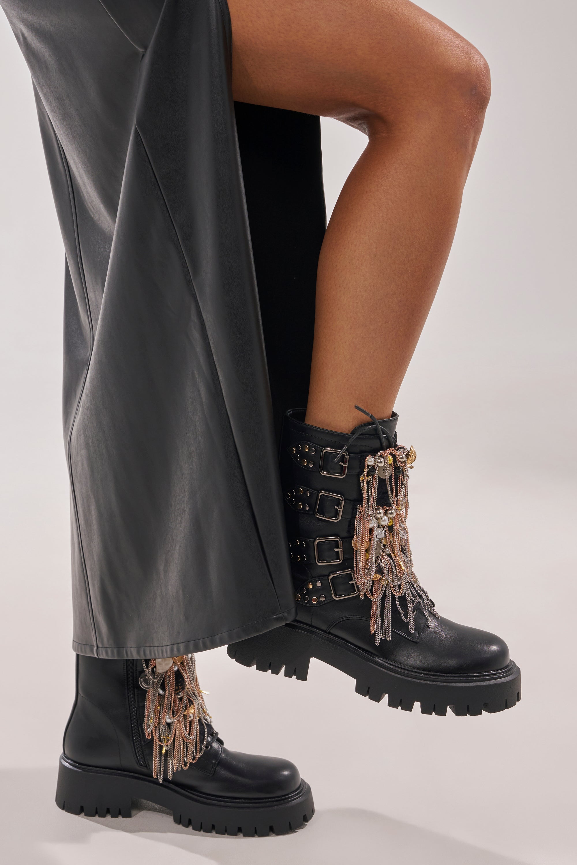 Wearing a long black leather skirt and AZALEA WANG MEGUMI BLACK METAL HARDWARE COMBAT BOOTIE with chunky heels, studs, and dangling chains, a person stands with one leg slightly raised. 3