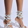 A person wearing the AZALEA WANG MEIER WHITE EMBELLISHED STILETTO SANDAL, featuring intricate, sparkling leaf-like designs on the straps. The sandals showcase a faux leather upper in white with iridescent embellishments and are photographed against a light background.