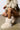 A person squats wearing a beige jacket with zip and cord details, paired with matching shorts. Their MELANIE BEIGE FURRY BOOTIE, soft and cream-colored, contrasts with the smooth, light brown floor. The minimalist setting is bright and open, enhancing the cozy yet chic vibe of their ensemble.