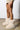 A person stands wearing beige, knee-length shorts and the chic MELANIE BEIGE FURRY BOOTIE with a flat sole. The floor is concrete, and the background is plain white.