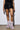 A person in a stylish, cold weather ensemble stands indoors. Their black outfit glimmers with metal accents, perfectly paired with the MELANIE PINK LEOPARD FURRY BOOTIE, featuring luxurious faux fur. Only their lower body is visible, highlighting the booties' pink and black leopard pattern from feet to mid-calf.