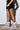 A person dressed in a black outfit stands against a white wall sporting the MELANIE PINK LEOPARD FURRY BOOTIE. The pink faux fur boots, adorned with unique black spots, cast dancing shadows on the wall, highlighting their texture and contributing a stylish touch perfect for colder weather.