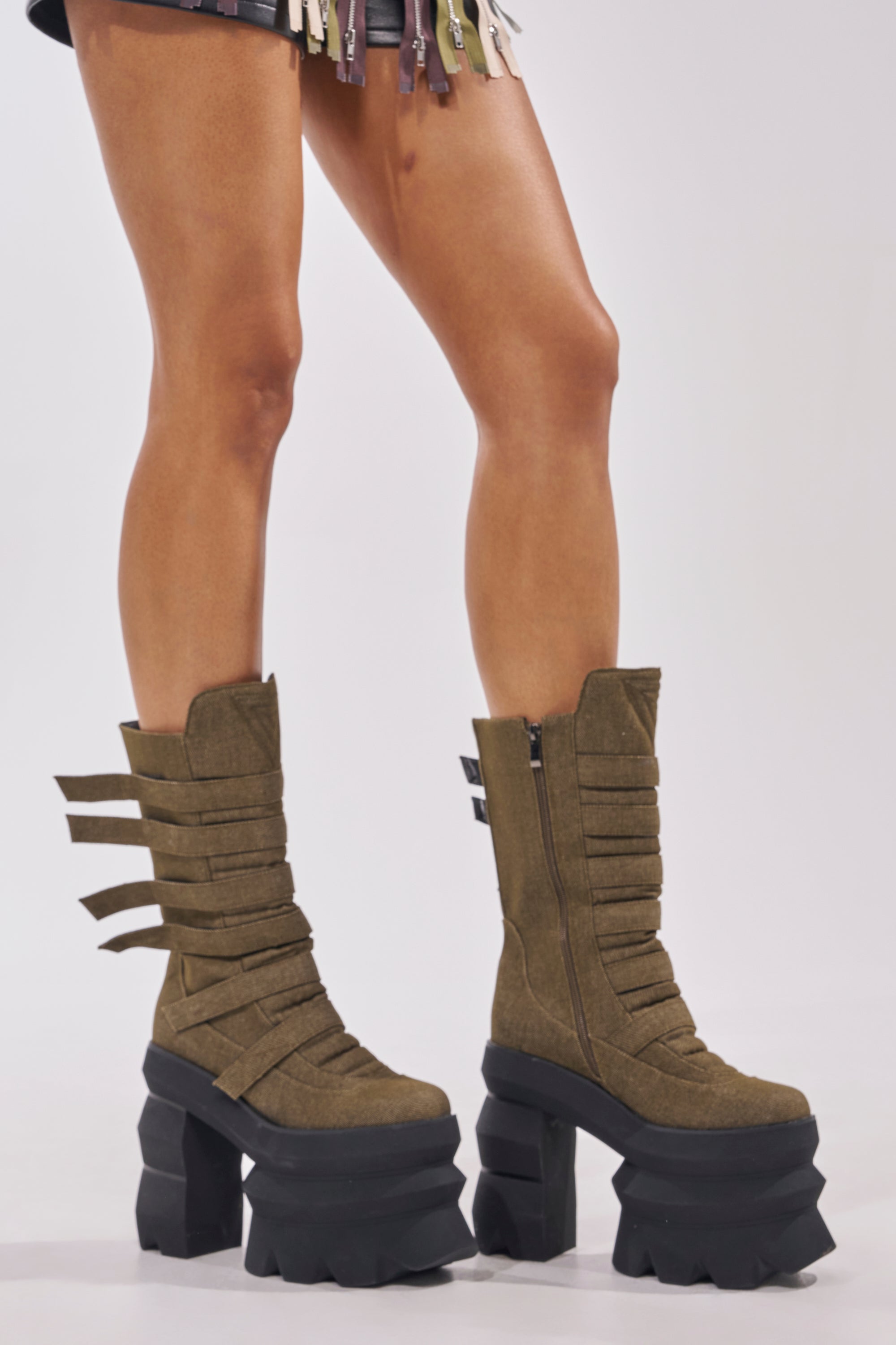 A person stands on a white background wearing the AKIRA GOLD MENACING OLIVE BLOCK PLATFORM BOOTIE, featuring multiple horizontal straps and chunky black platform heels. Part of their skirt with hanging strips is also visible. 1