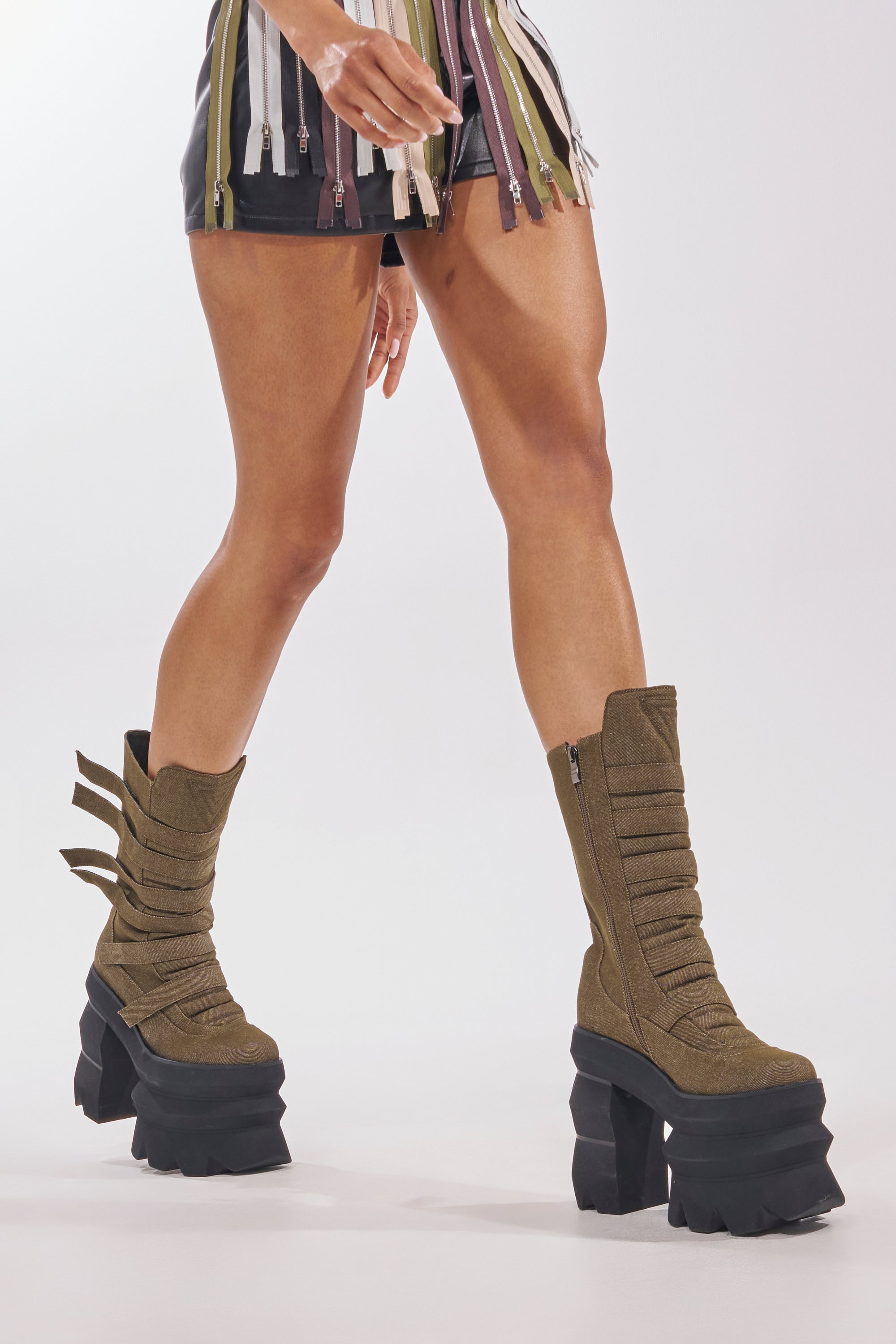 A person wearing the AKIRA GOLD MENACING OLIVE BLOCK PLATFORM BOOTIE stands against a plain white background, showing only their lower half in a short multicolored fringe outfit. 2