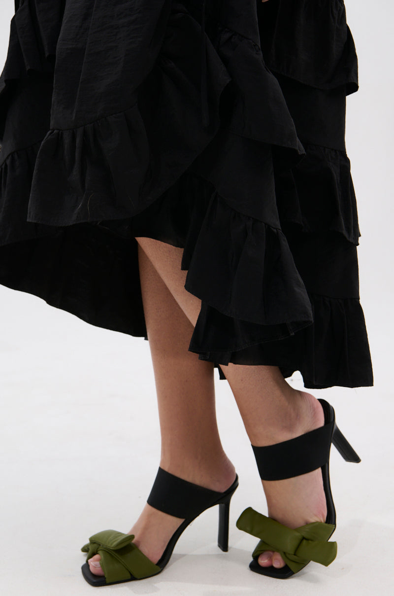 A person in a black ruffled skirt and AZALEA WANG MENES BLACK PUFF DETAIL SANDAL, featuring rectangular stiletto heels, black straps, and green bows, stands on a light-colored surface.