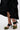 A person in a black ruffled skirt and AZALEA WANG MENES BLACK PUFF DETAIL SANDAL, featuring rectangular stiletto heels, black straps, and green bows, stands on a light-colored surface.