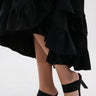 A person in a black ruffled skirt and AZALEA WANG MENES BLACK PUFF DETAIL SANDAL, featuring rectangular stiletto heels, black straps, and green bows, stands on a light-colored surface.