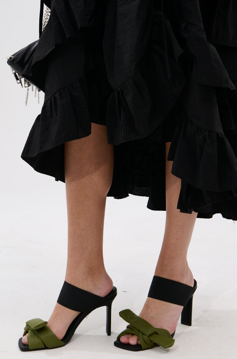Wearing a black ruffled skirt and AZALEA WANG MENES BLACK PUFF DETAIL SANDAL with green bows and a rectangular stiletto heel, the person stands on a white surface with a small black bag featuring silver hardware for a chic look.