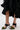 Wearing a black ruffled skirt and AZALEA WANG MENES BLACK PUFF DETAIL SANDAL with green bows and a rectangular stiletto heel, the person stands on a white surface with a small black bag featuring silver hardware for a chic look.
