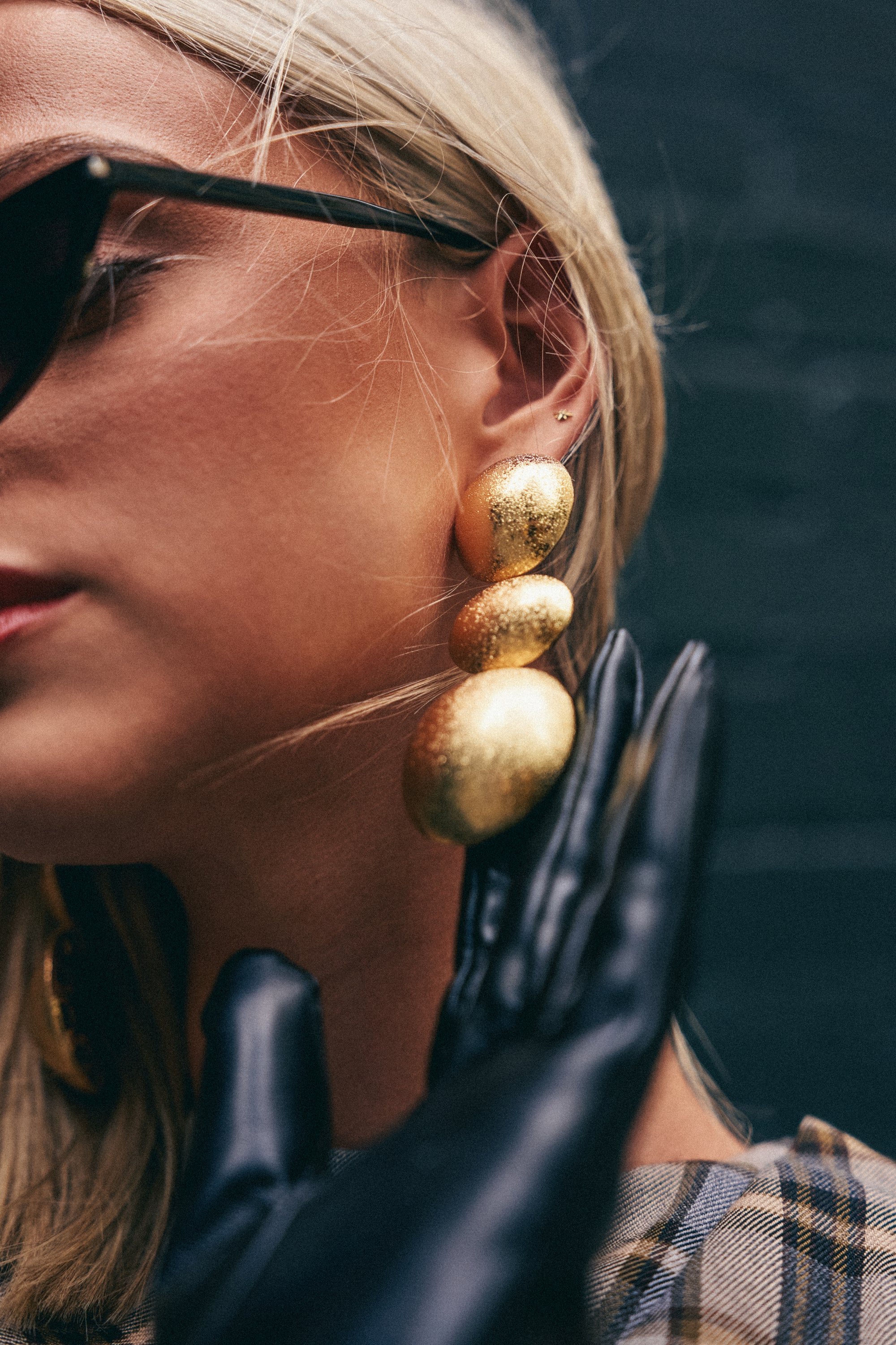 A woman with blonde hair wears black sunglasses and black gloves, holding her gloved hand near her face. She is styled in a plaid-patterned garment and showcases the BALLS TO THE WALL EARRING IN GOLD.
