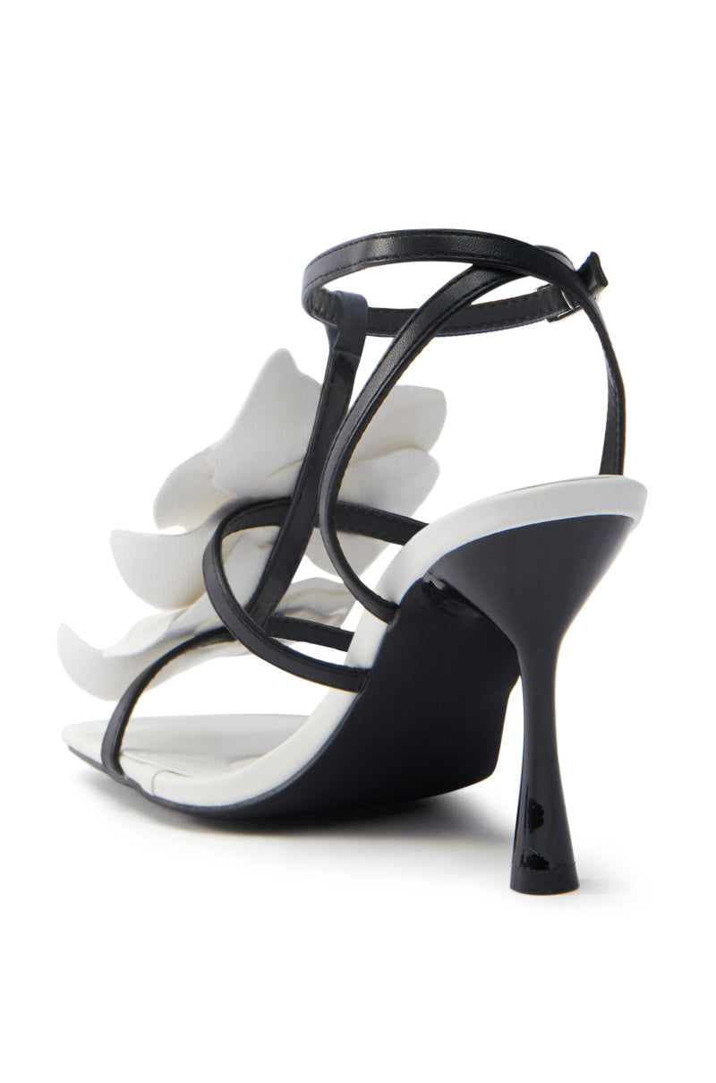 The AZALEA WANG MERCI BLACK AND WHITE FLORAL STILETTO SANDAL is a vegan leather high-heeled sandal featuring thin black straps, an open toe design with decorative white ruffle straps, and a glossy black stiletto heel with a slight base flare. 5