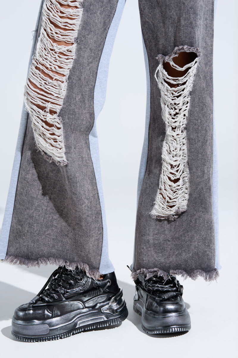 A close-up of a person's legs in distressed, ripped gray jeans with frayed edges, paired with KOI FOOTWEAR METAMORPHOSIS BLACK FLAT DISTRESSED SNEAKERS, standing on a light surface. 0