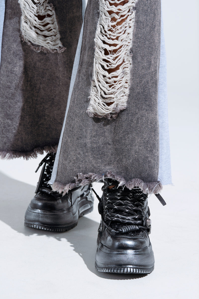 Close-up of a person wearing KOI FOOTWEAR METAMORPHOSIS Black Flat Distressed Sneakers with wide-leg, heavily ripped pants. The light-colored floor contrasts with the blurred background. 1