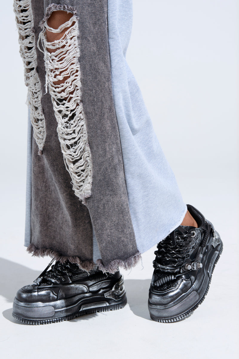 A close-up of a person in distressed, two-tone jeans with large rips on one leg, styled with KOI FOOTWEAR METAMORPHOSIS BLACK FLAT DISTRESSED SNEAKER. The backdrop is plain and light-colored. 6