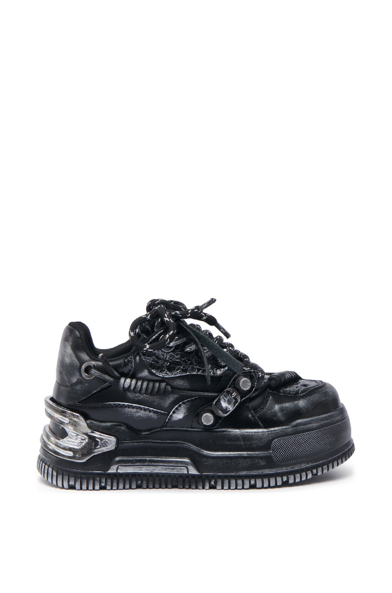 The KOI Footwear Metamorphosis Black Flat Distressed Sneaker features a chunky, exaggerated sole, metallic accents, thick laces, and layered textures, embodying bold industrial streetwear style against a plain white background. 2