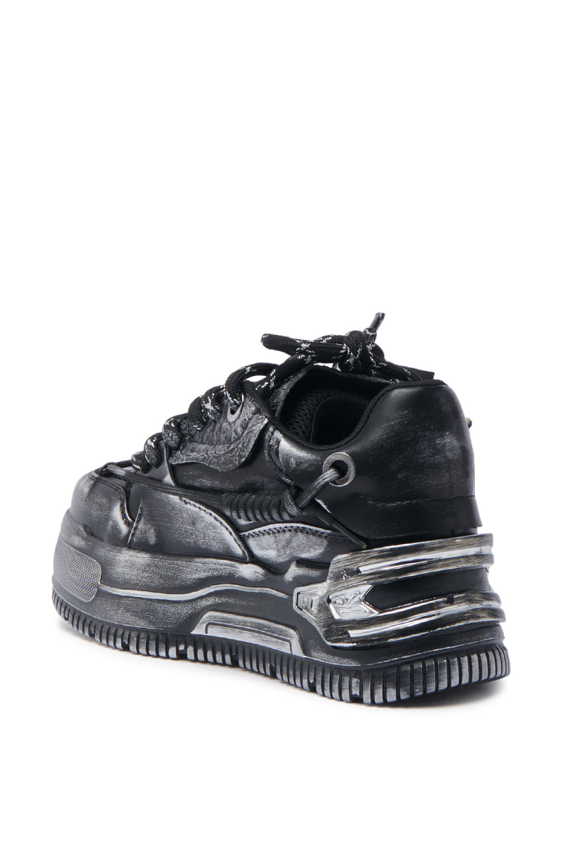 The KOI FOOTWEAR METAMORPHOSIS BLACK FLAT DISTRESSED SNEAKER is a black flatform sneaker with thick, chunky soles, metallic silver heel accents, textured detailing, black laces, and a bold futuristic design. Viewed from the back left. 4