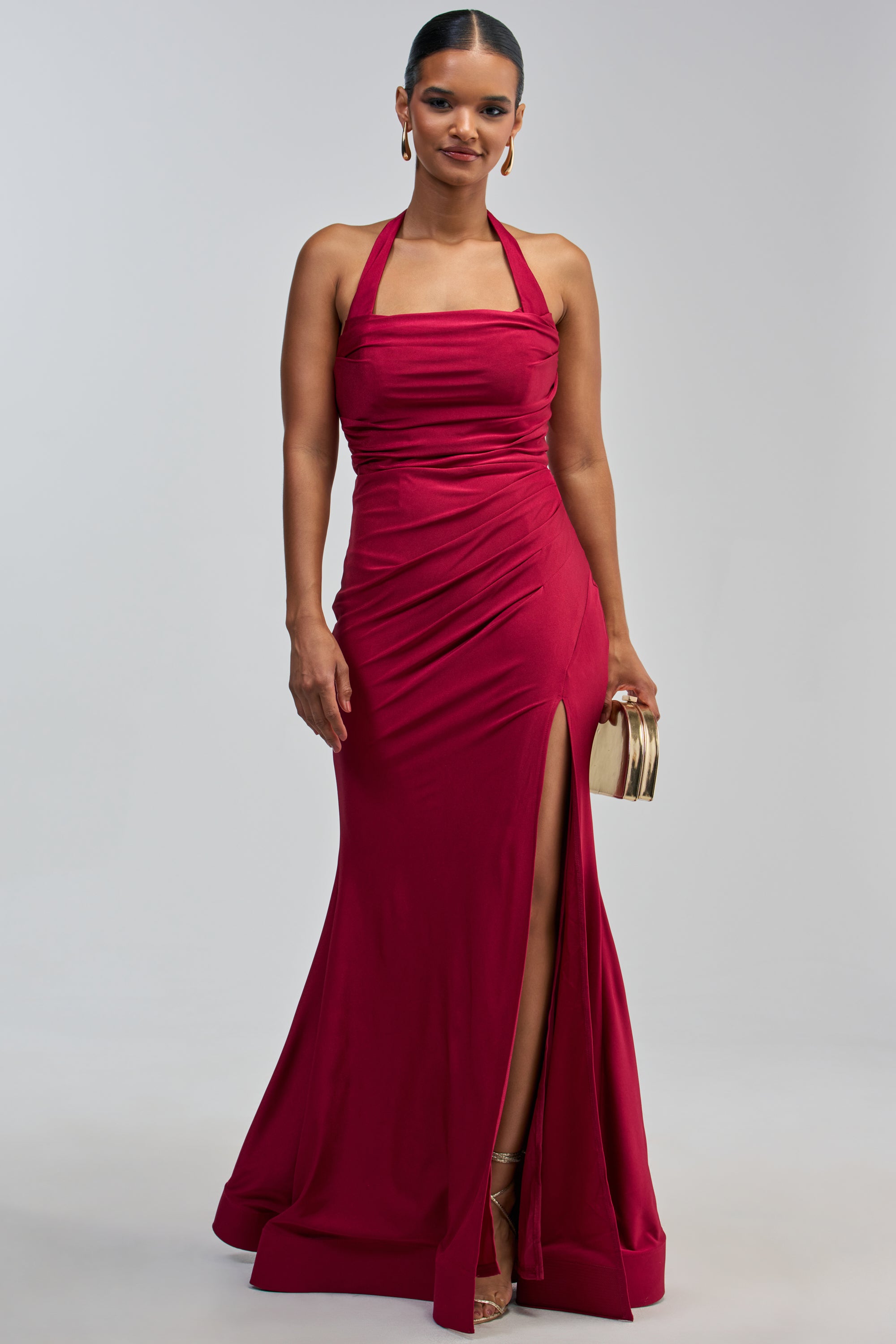 A woman stands against a plain light background wearing the MYSTERIOUS MUSE HIGH SLIT MAXI DRESS—a sleeveless, floor-length burgundy gown with a halter neckline and high slit. She accessorizes with gold hoop earrings, a gold clutch, and heels.