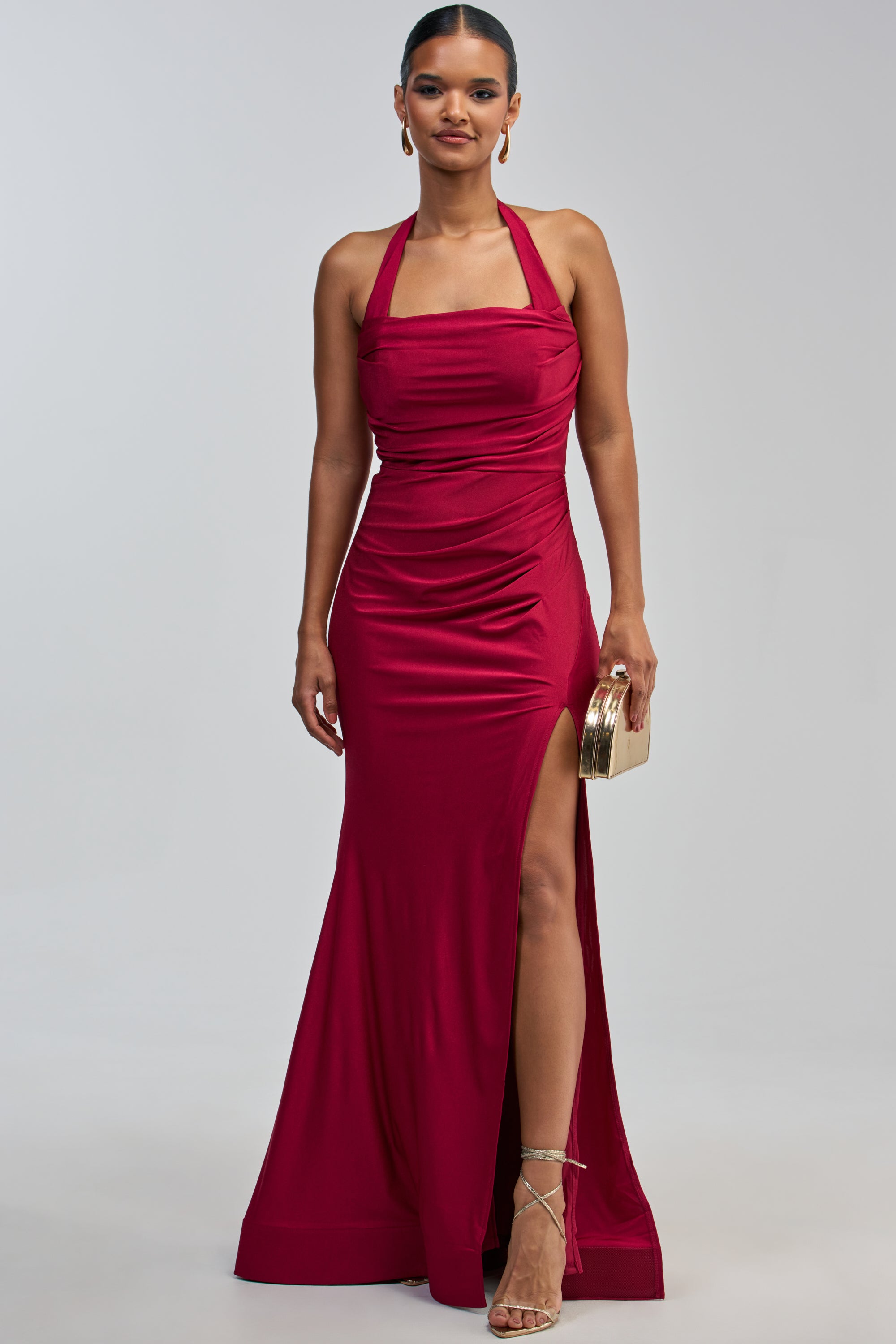 A woman stands against a plain background wearing the MYSTERIOUS MUSE HIGH SLIT MAXI DRESS—a sleeveless, floor-length burgundy maxi dress featuring a halter neckline and thigh-high slit. She accessorizes with a gold clutch and strappy heels.