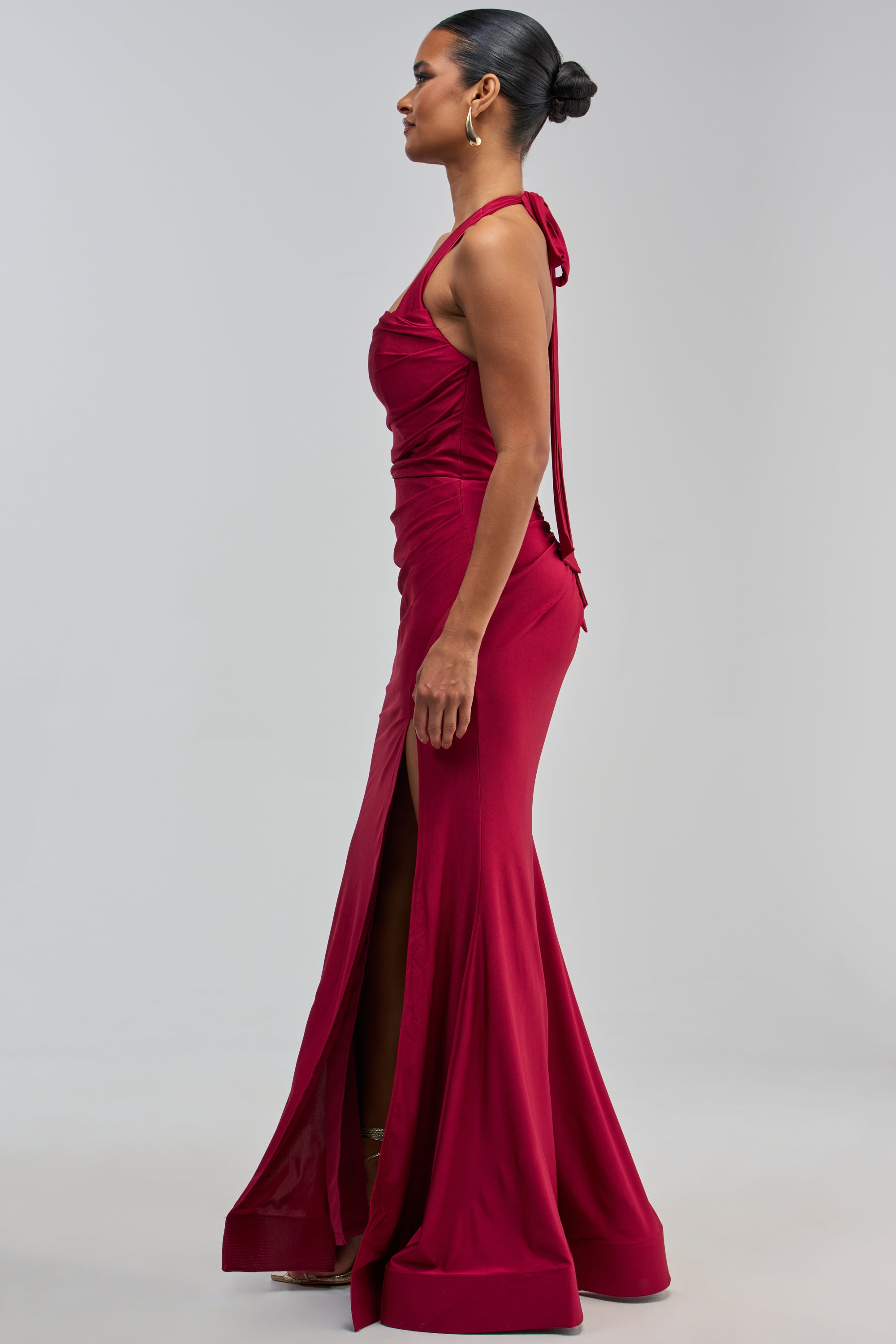A woman stands in profile against a plain light background, wearing the MYSTERIOUS MUSE HIGH SLIT MAXI DRESS—a sleeveless, floor-length burgundy gown featuring a striking high slit. Her hair is styled in a sleek bun, complemented by gold hoop earrings, creating an elegant look ideal for a chic wedding guest.
