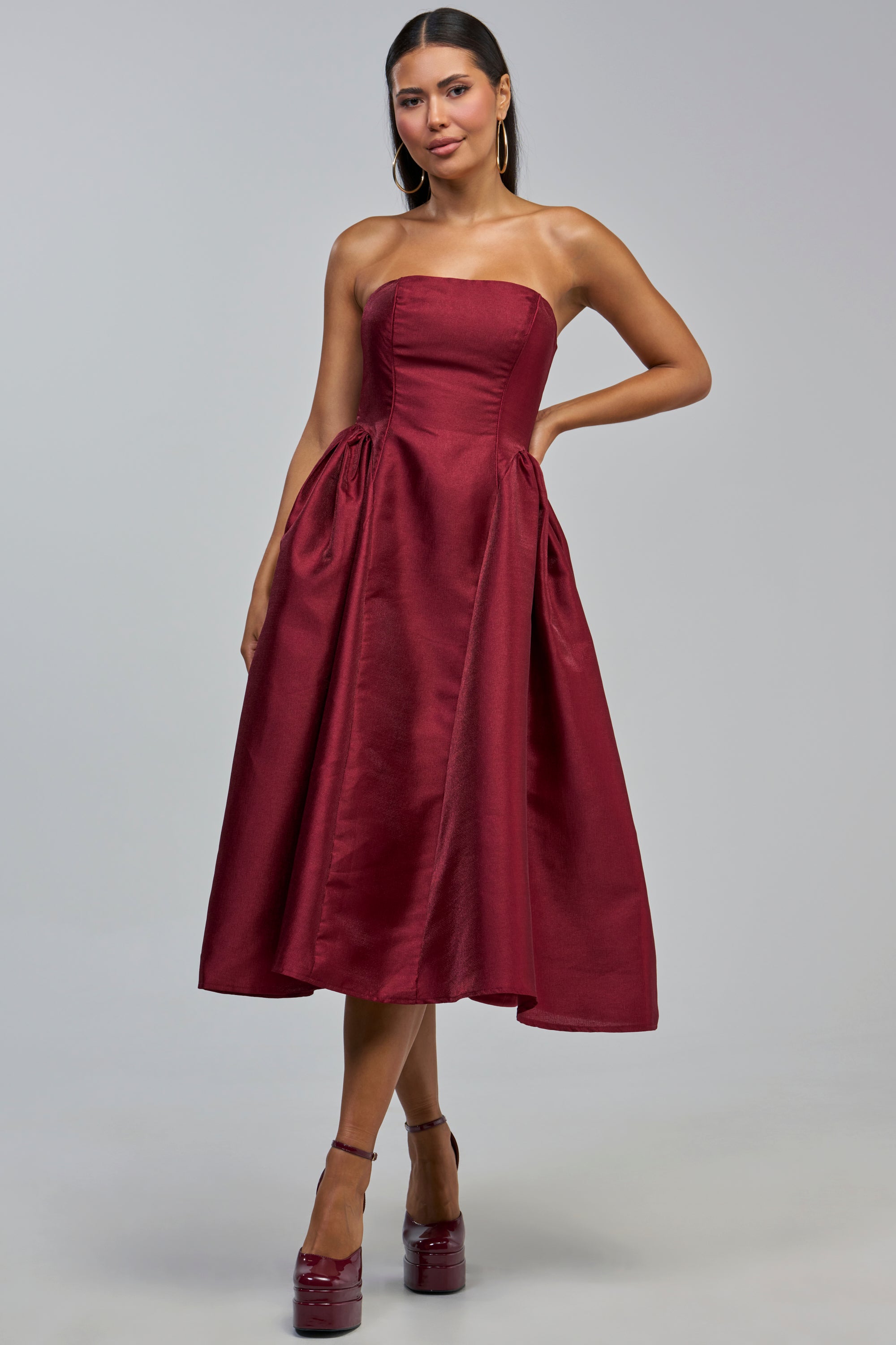 A woman poses with one hand on her hip, wearing the ALWAYS DANCING STRAPLESS MIDI DRESS IN WINE—a strapless, A-line midi dress crafted from shiny fabric. Paired with matching high-heeled shoes and hoop earrings, she stands against a plain gray background—an elegant option for cocktail parties or as a wedding guest dress.