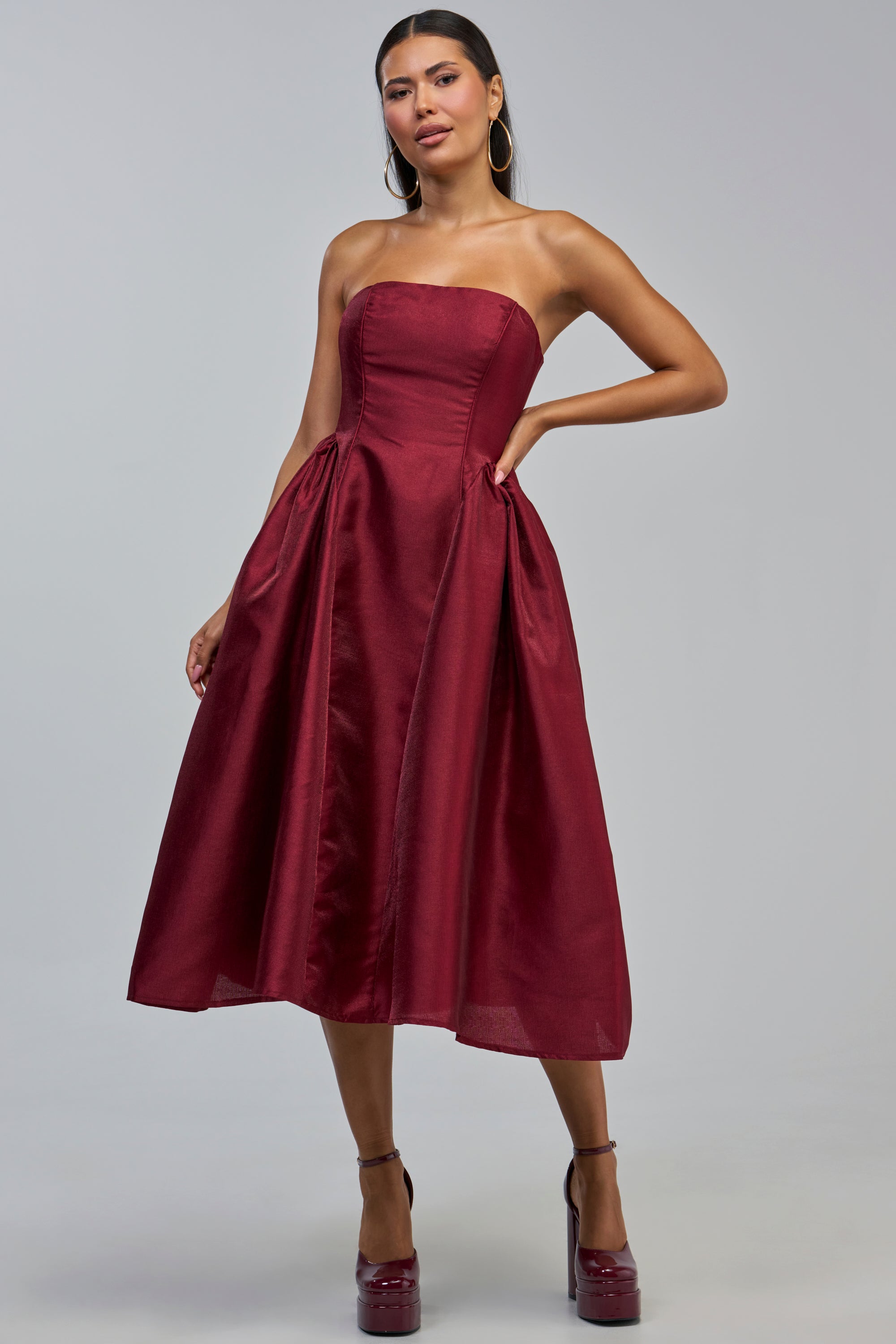 Model showcases the ALWAYS DANCING STRAPLESS MIDI DRESS IN WINE, featuring a fitted bodice and flared skirt in a rich burgundy hue. Styled as an elegant cocktail party or wedding guest dress, she poses with one hand on her hip, accessorized with platform heels and hoop earrings.