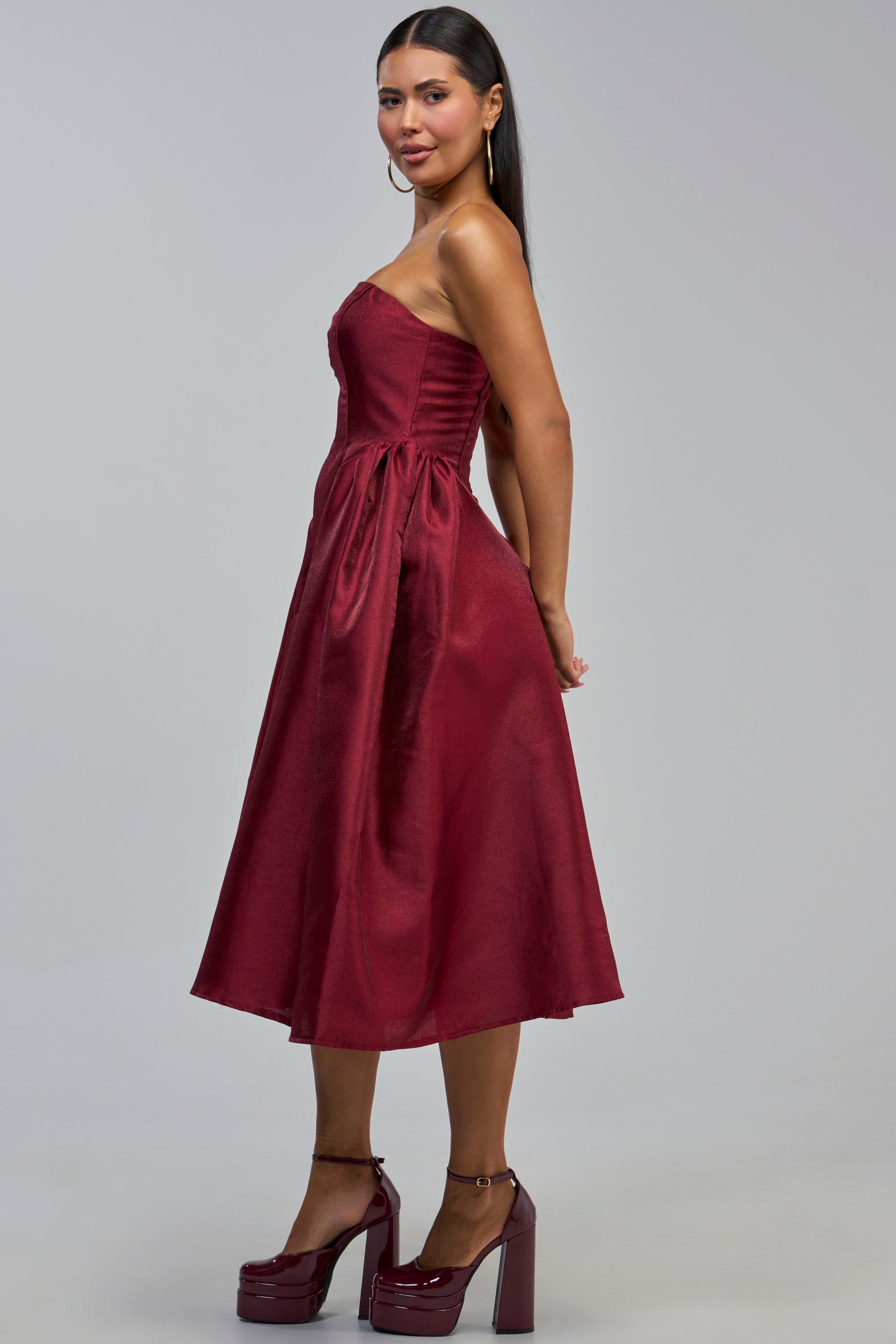 A woman with long dark hair, hoop earrings, and platform heels stands sideways against a plain gray background, showcasing the ALWAYS DANCING STRAPLESS MIDI DRESS IN WINE. This burgundy dress features a fitted bodice and flared skirt, making it an ideal choice for a wedding guest or cocktail party look.