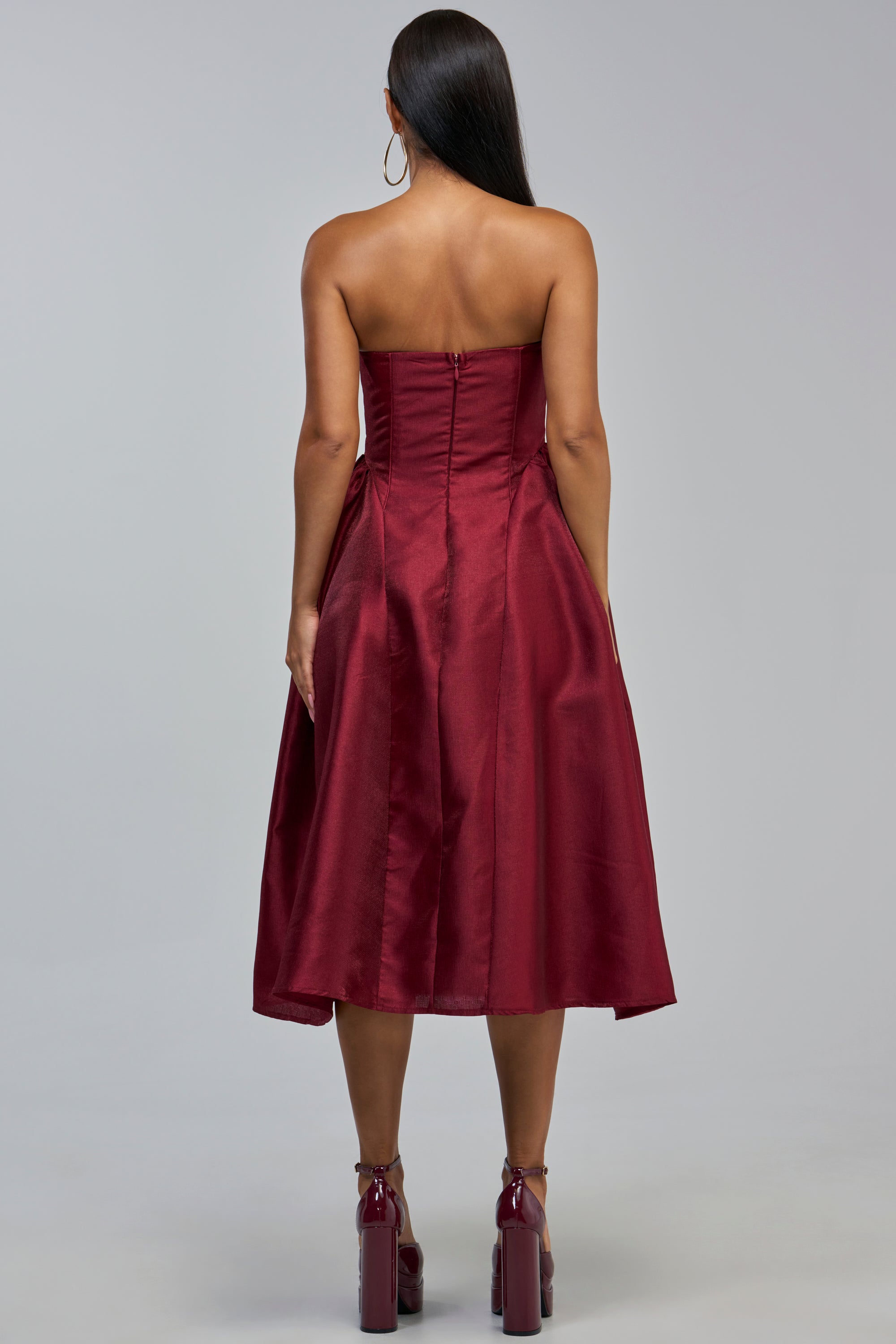 A woman with long straight hair, seen from behind, wears the ALWAYS DANCING STRAPLESS MIDI DRESS IN WINE—an ideal choice for a chic cocktail party or as an elegant wedding guest dress—paired with matching high-heeled shoes. She stands against a plain grey background.