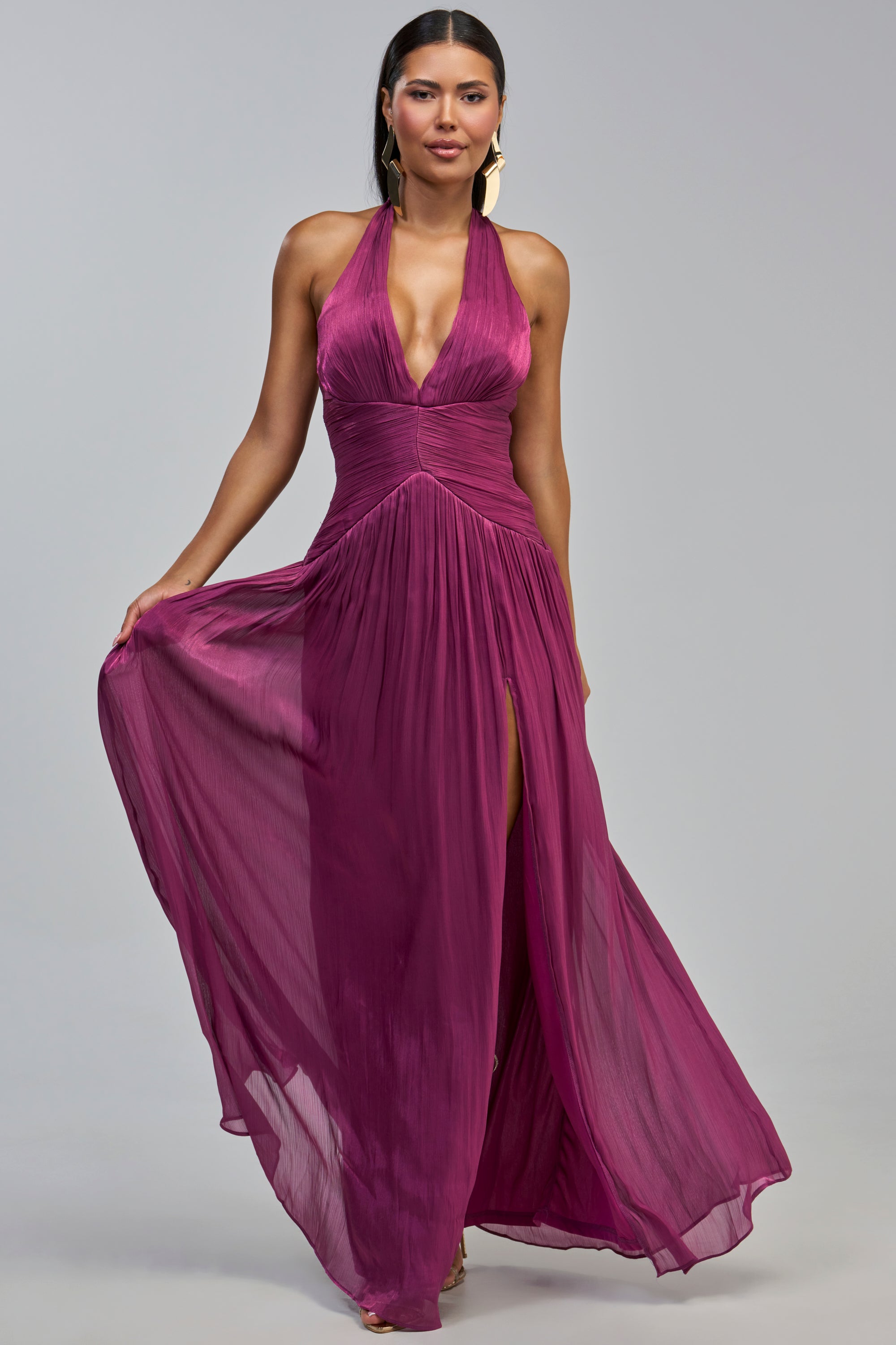 A woman with long, straight hair stands against a plain gray background, holding out one side of the flowing skirt of the TOO STYLED TO CARE MAXI DRESS IN PLUM. This sleeveless maxi dress features a deep V-neckline, halter design, and high slit, showcasing a graceful silhouette that’s perfect as a wedding guest dress.
