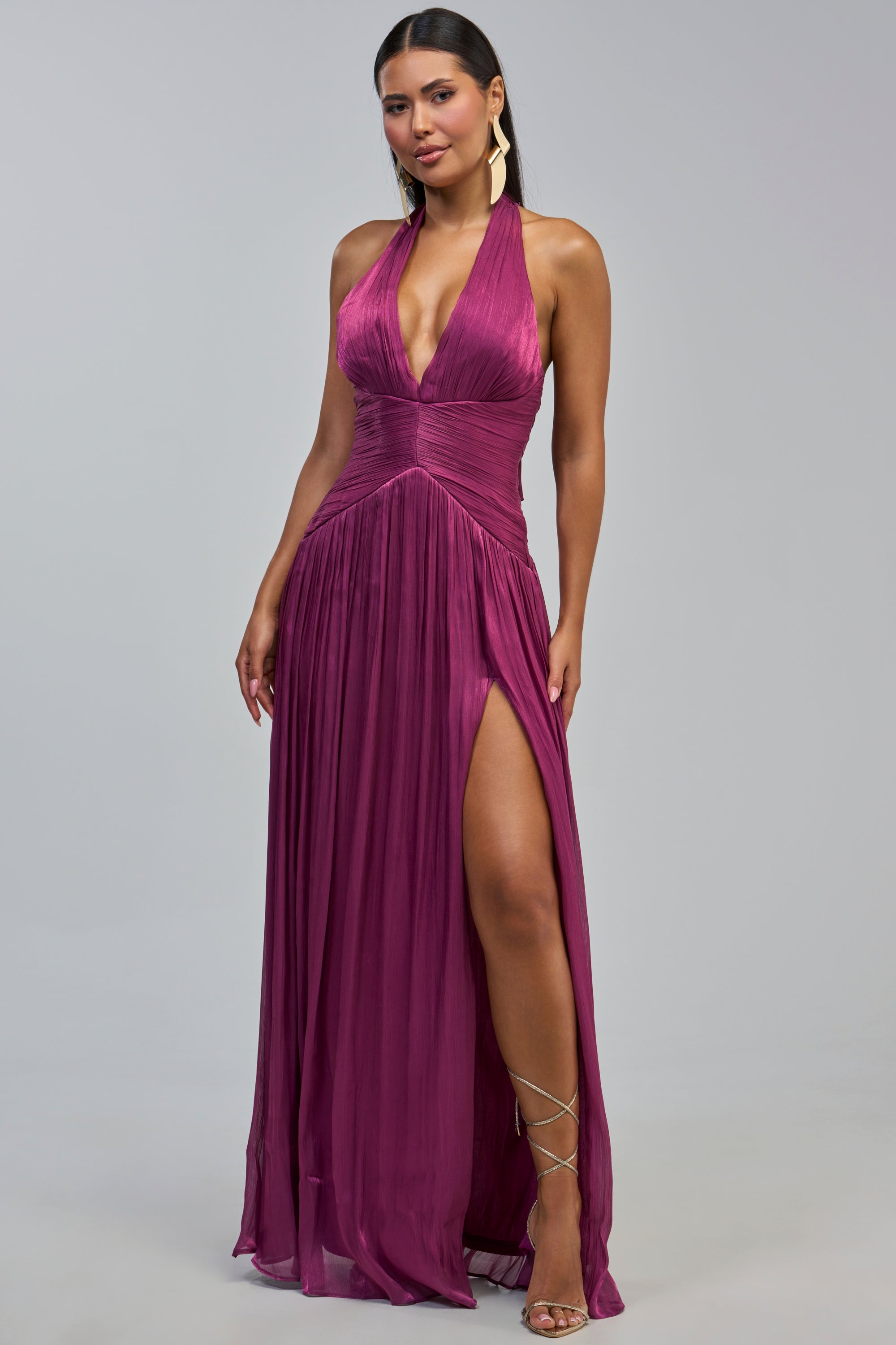 A woman models the TOO STYLED TO CARE MAXI DRESS IN PLUM—a sleeveless gown with a deep v-neckline and high slit—standing against a plain light background. Perfect as a wedding guest dress, her look is completed with long dark hair, gold earrings, and strappy heels.