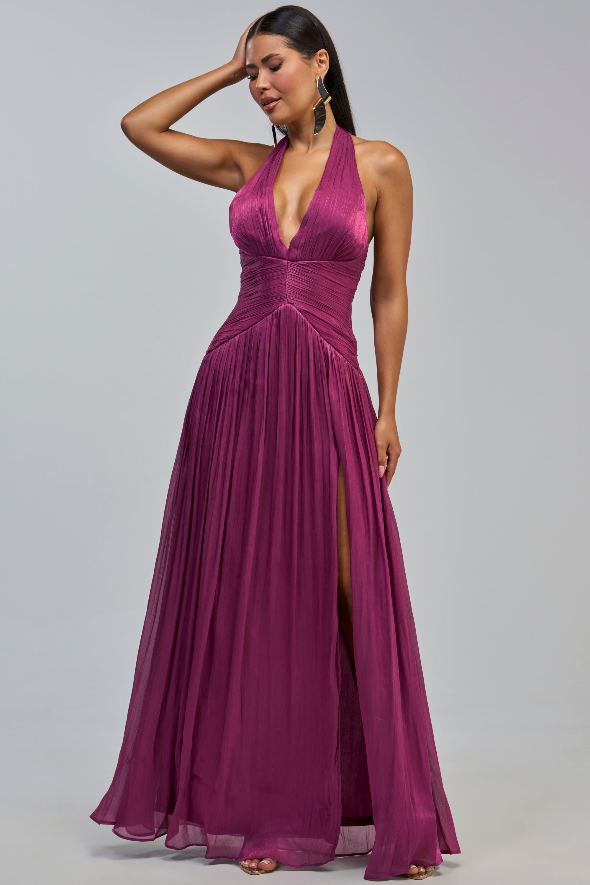 A woman poses in the TOO STYLED TO CARE MAXI DRESS IN PLUM, featuring a sleeveless halter neckline, deep plum hue, plunging front, fitted waist, flowing pleated skirt, and high slit. With long dark hair and statement earrings, she rests one hand on her head—showcasing this stunning dress as the ideal choice for any elegant wedding guest occasion.
