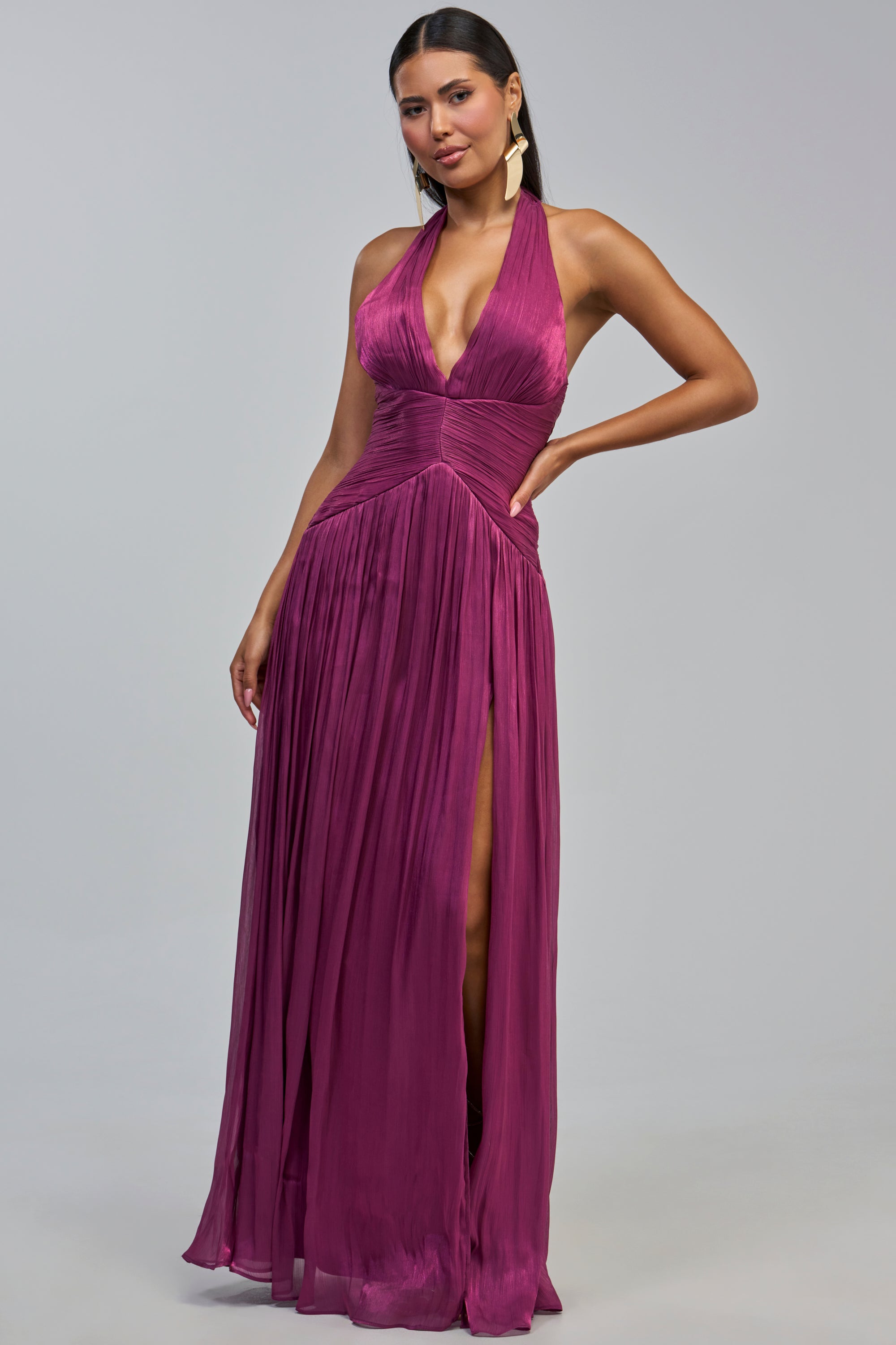 A woman with long dark hair and gold earrings stands against a plain light gray background, wearing the TOO STYLED TO CARE MAXI DRESS IN PLUM—a sleeveless, deep v-neck, pleated chiffon maxi dress with a high slit—making for an elegant wedding guest look.