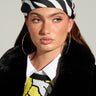 A woman styled with the ZEBRA HEADSCARF and large hoop earrings gazes to the side. She wears a black faux fur coat over a sequined yellow top with a white collar. The simple light gray backdrop accentuates her bold hair accessory choice.